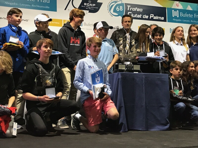 A group of young people stand and kneel on a stage during an awards ceremony, some holding plaques and trophies, with sailboat-themed banners and a large silver trophy behind them.