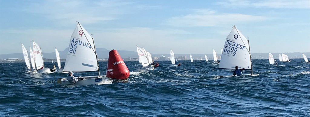 A group of small sailboats race around a large red buoy on choppy blue water, with distant hills and a city skyline visible under a partly cloudy sky.