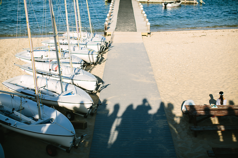 Several small sailboats are lined up on a sandy beach beside a wooden dock extending into the water, with shadows of people cast on the walkway and two people sitting at a picnic table nearby.