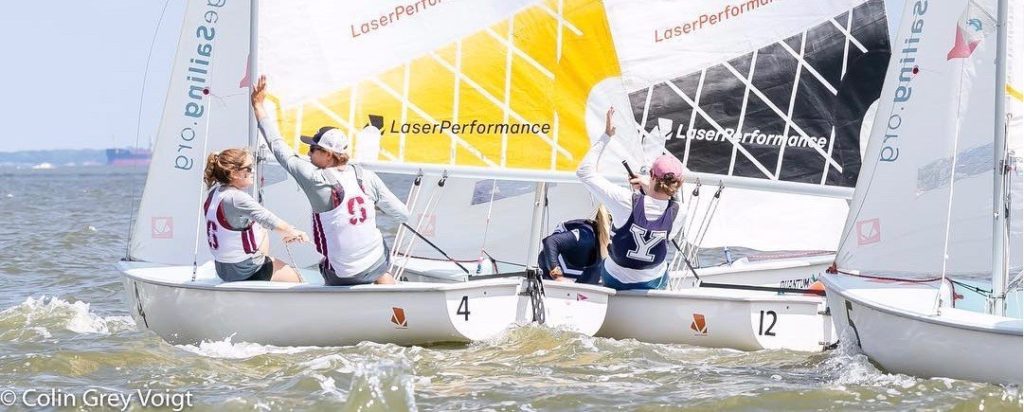 Two teams of sailors in numbered sailboats high-five each other on the water during a regatta. The sailors wear life jackets and caps, and “LaserPerformance” is visible on the sails.