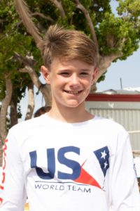 A smiling boy with light brown hair stands outdoors, wearing a white shirt that says "USA WORLD TEAM" with a stylized flag design. Green trees and a metal building are visible in the background.