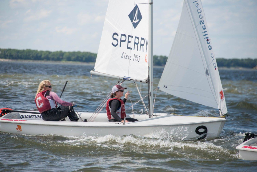 Two people wearing life jackets sail a small white boat with the number 9 and a “SPERRY” sail on choppy water. Another sailboat is partially visible behind them. Trees line the distant shore under a partly cloudy sky.