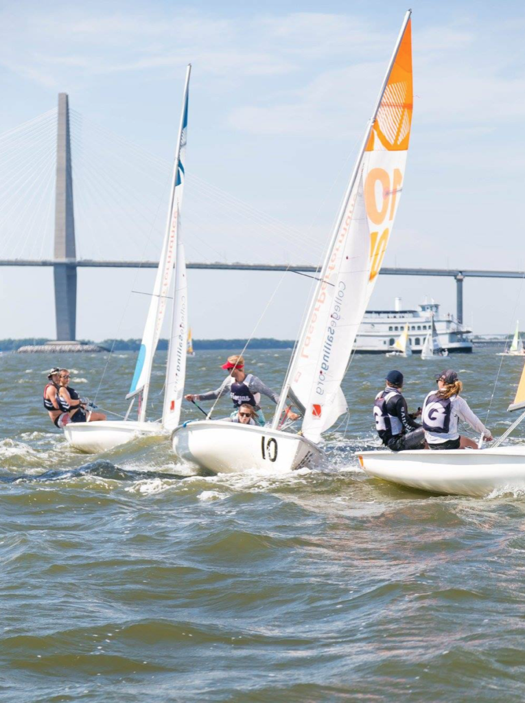 Three small sailboats with teams of two race on choppy water near a bridge, with a large boat and clear blue sky in the background. The sails display numbers and logos.