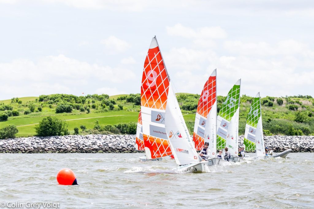 Four small sailboats with colorful orange and green sails race on a lake near a red buoy, with green hills and a rocky shoreline in the background under a cloudy sky.