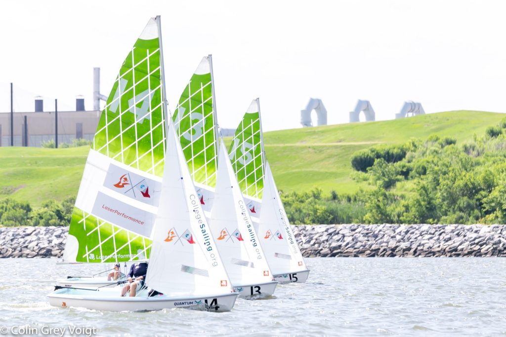 Three sailboats with bright green and white patterned sails race on the water near a rocky shoreline, with grassy hills and industrial buildings in the background.