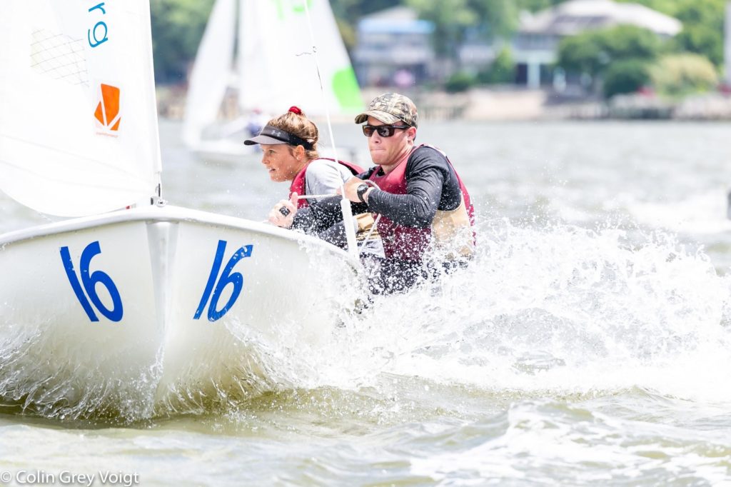 Two people wearing caps and life jackets are sailing a small boat marked with the number 16, cutting through choppy water with splashes, with blurred sailboats and shoreline in the background.
