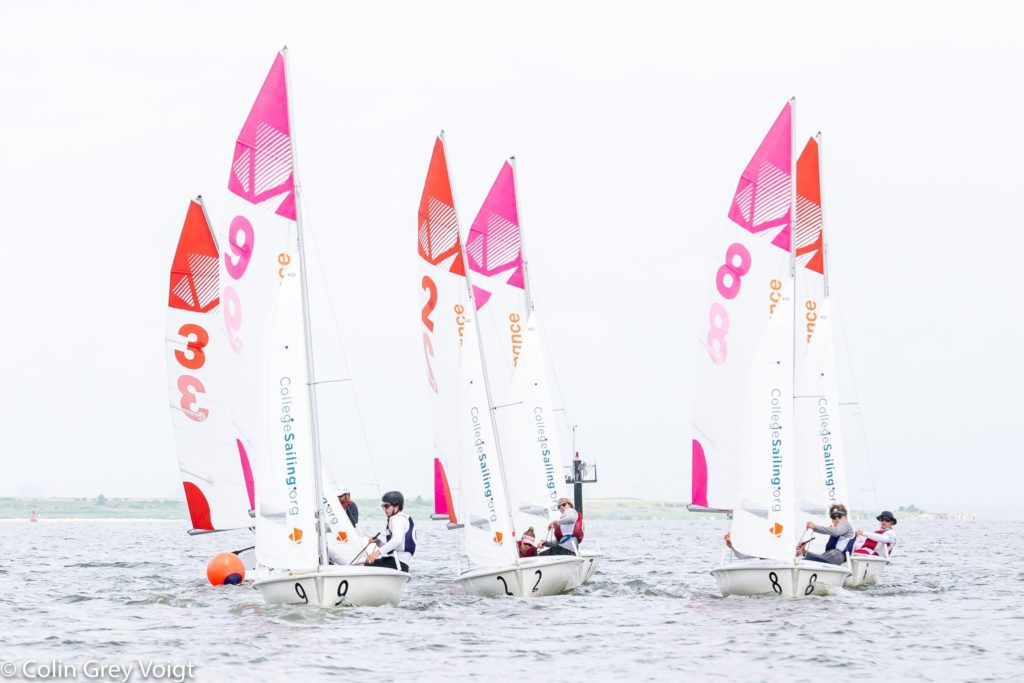 Four small sailboats with pink and orange sails, each carrying two people, race on a calm body of water. The sails display large numbers, and the boats are labeled 0, 2, 7, and 8. The sky is overcast.