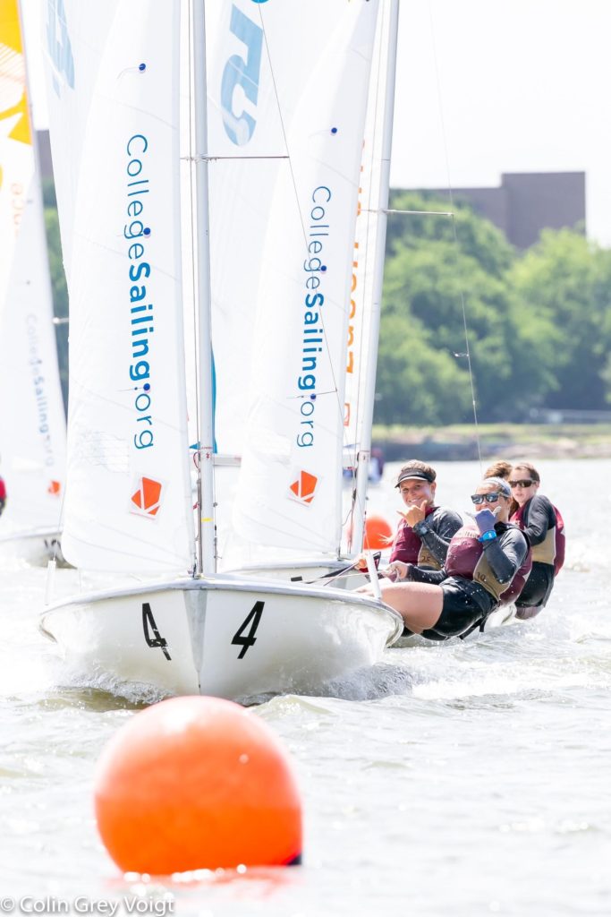 Two teams of sailors compete in small sailboats on a sunny day, leaning out over the water. Orange buoys mark the course, and "collegesailing.org" is visible on the sails. Trees and buildings are in the background.