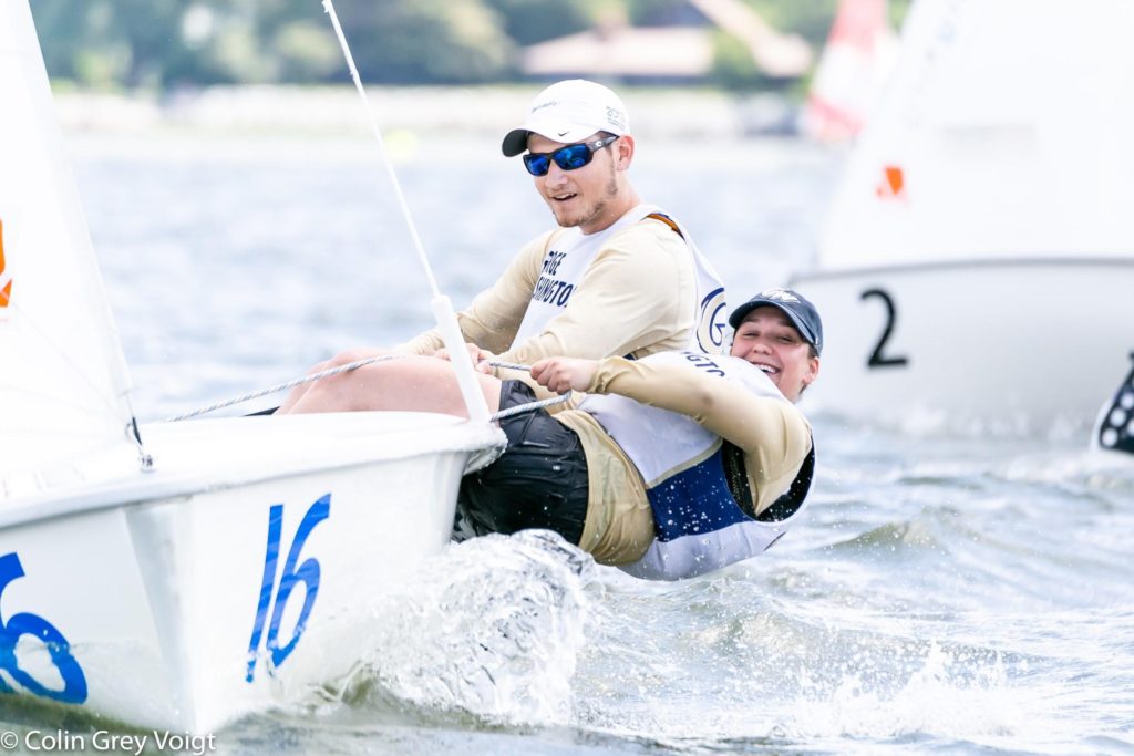 Two sailors wearing sunglasses and life vests lean out of a small sailboat numbered 16 as they race in choppy water, with another boat numbered 2 visible in the background.