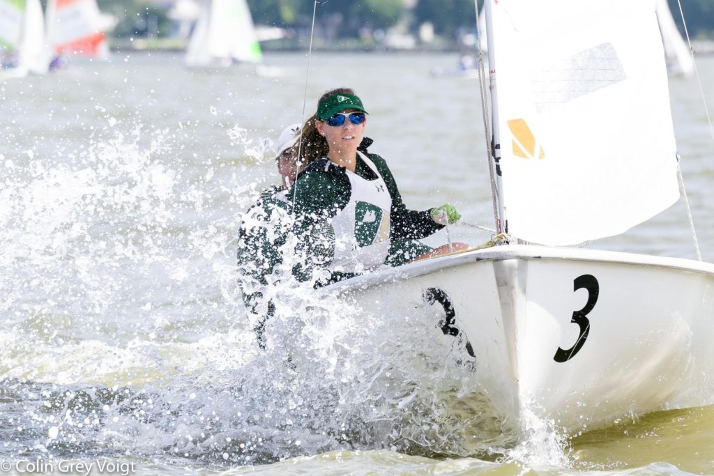 A sailor wearing sunglasses and a cap steers a sailboat marked with the number 3, as water splashes up around the bow. Other sailboats and trees are visible in the blurred background.
