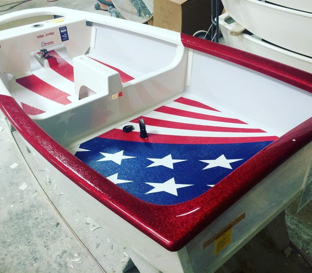 Small boat with a red glitter border and an American flag design on the interior floor, featuring white stars on a blue background and red and white stripes. The boat is indoors, surrounded by other boats and workshop materials.