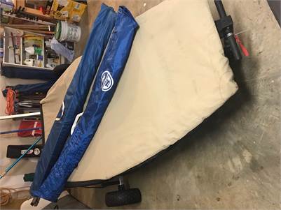 A folding cot with beige fabric and blue trim is set up on a concrete floor in a garage, surrounded by various tools and storage items. The cot has wheels attached for easy transport.