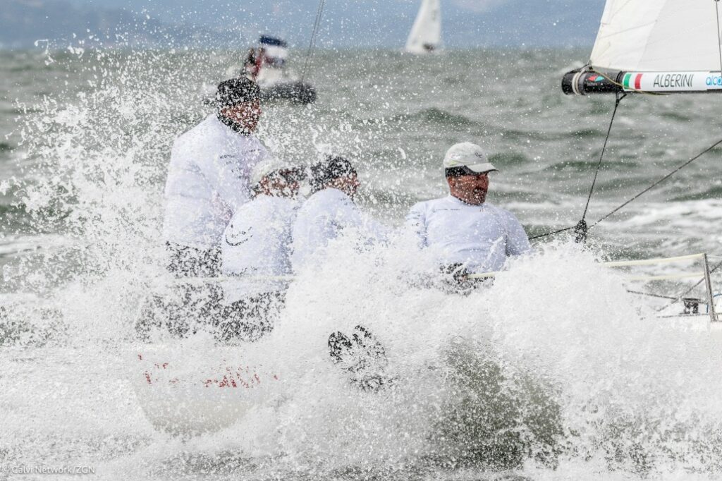 Four sailors in white jackets are splashed by a large wave while racing on a sailboat in choppy waters, with another boat visible in the background.