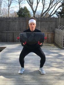 A woman in athletic clothing and a white headband is holding a sandbag at chest level while performing a squat on a wooden deck outdoors. Leafless trees and a wooden fence are in the background.