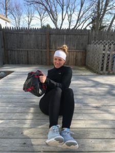 A woman in athletic wear sits on a wooden deck outdoors, holding a weighted sandbag and smiling. Leafless trees and a wooden fence are visible in the background on a sunny day.
