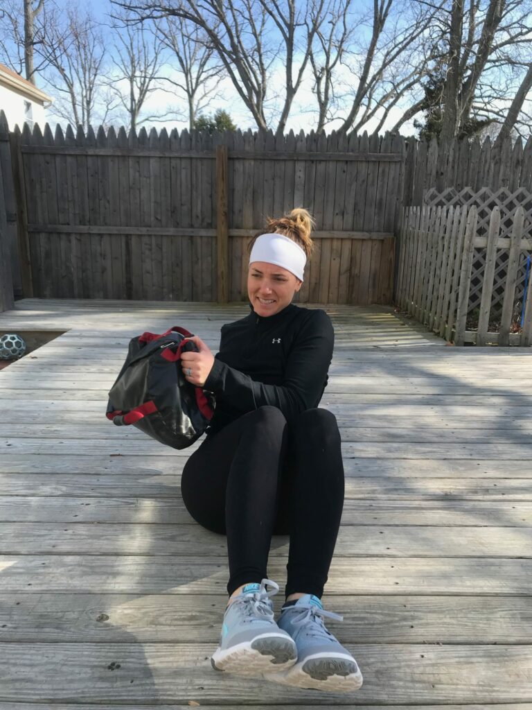 A woman in athletic wear sits on a wooden deck outdoors, holding a weighted sandbag and smiling. Leafless trees and a wooden fence are visible in the background on a sunny day.