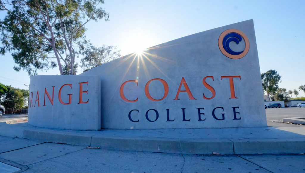 A large concrete sign reads 'Orange Coast College' in orange and blue letters, with a wave logo. The sun shines behind the sign, and trees and a parking lot are visible in the background.