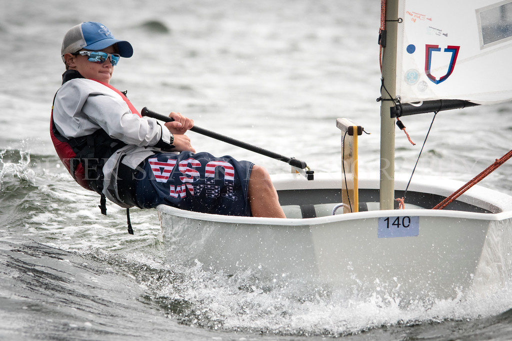 A young sailor in sunglasses and a life jacket leans back in a small sailboat on choppy water, with "USA" printed on his shorts and a blue cap on his head.