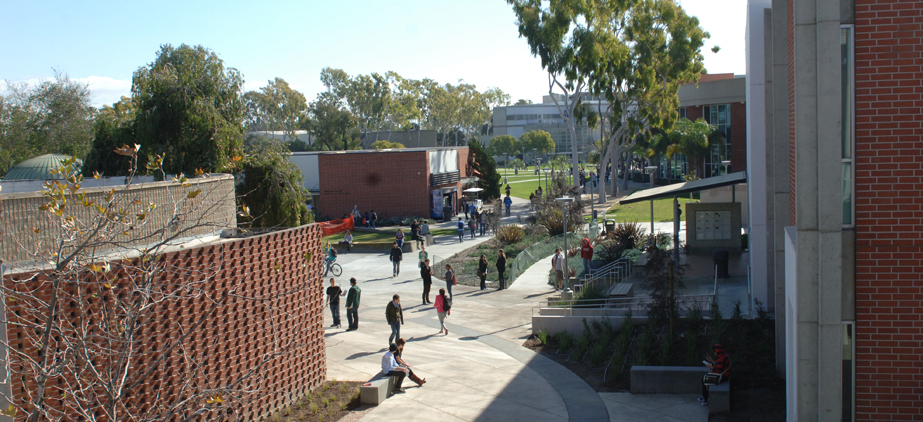 A college campus scene with students walking, sitting, and biking along a wide pathway bordered by brick buildings, trees, and landscaped greenery on a sunny day.