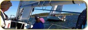 People are sailing on a boat in open water, approaching another sailboat with "BLACK ROCK" on its sail. The weather is clear and sunny, and a green shoreline is visible in the background.