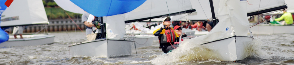 Sailboats with crews race closely on a choppy body of water, with white sails billowing and participants wearing life jackets and helmets. A shoreline with greenery is visible in the background.