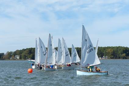 Several small sailboats with white sails race on a lake near a buoy, with wooded shoreline and a few houses visible in the background under a partly cloudy sky.