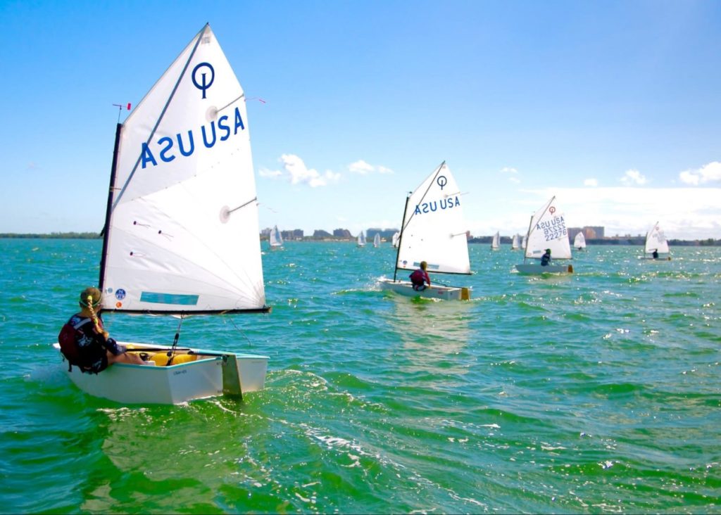 Several small sailboats, each with a single sailor, glide across green-blue water under a clear blue sky. The closest boat has "USA" on its sail. A distant shoreline is visible in the background.