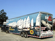 A large trailer decorated with images of sailboats and logos, attached to a black pickup truck, is parked on gravel with trees and power lines in the background.