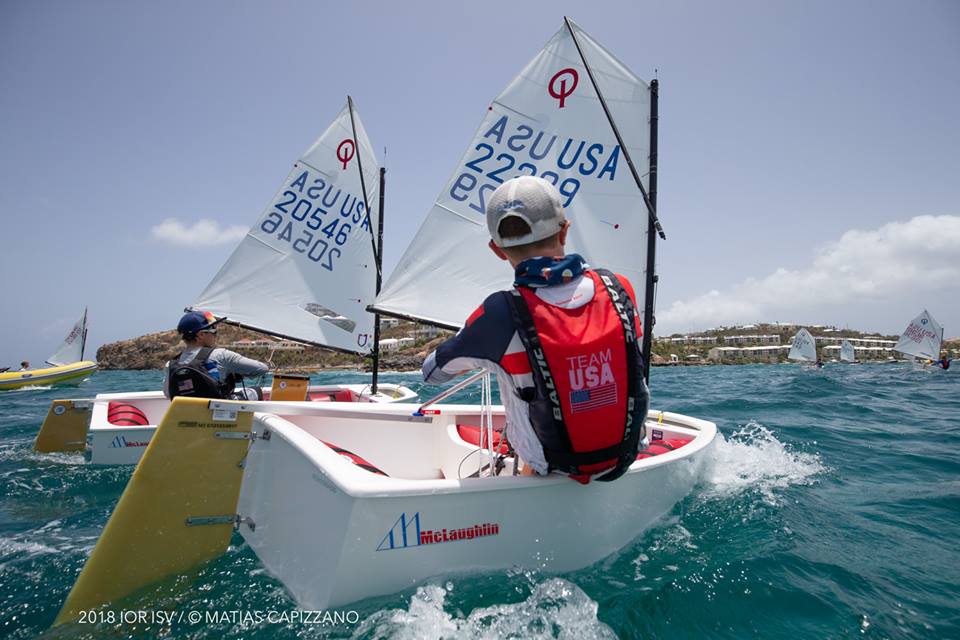 A young sailor wearing a 'Team USA' life vest navigates a small sailboat in a regatta, with other youth boats nearby on clear blue water; buildings and hills are visible in the background under a partly cloudy sky.