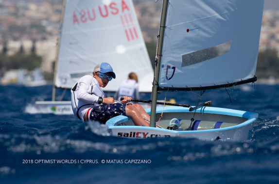A young sailor wearing sunglasses and a cap competes in a small sailboat on choppy blue water, with another sailboat and a blurred city in the background. Text at the bottom credits the 2018 Optimist Worlds in Cyprus.