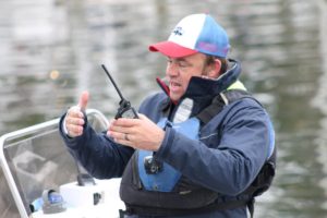 A man in a blue jacket and life vest sits on a boat, holding and speaking into a handheld radio, gesturing with his other hand over the water.