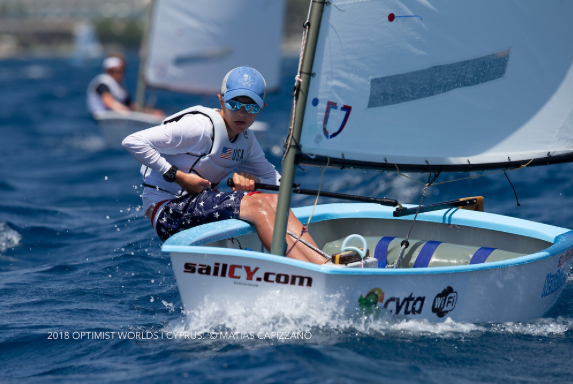 A young sailor wearing sunglasses and a cap navigates a small sailboat in a race on blue water, with other competitors in the background. The boat has "sailCY.com" printed on the side.