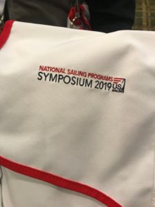 A white fabric bag or shirt with red trim displays the logo and text: "National Sailing Programs Symposium 2019, US Sailing," stitched in red and black.