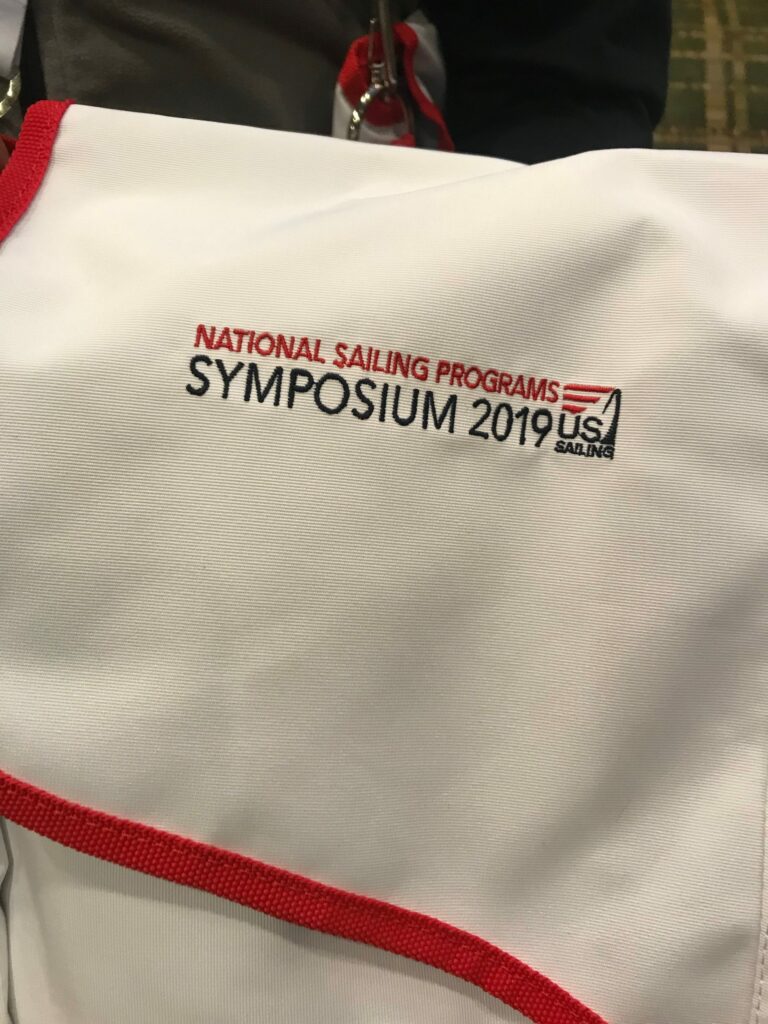 A white fabric bag or shirt with red trim displays the logo and text: 'National Sailing Programs Symposium 2019, US Sailing,' stitched in red and black.