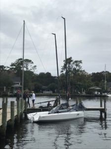 A group of people stands on a dock next to a small white sailboat with a tall mast on a calm, overcast day. Trees and houses are visible in the background across the water.