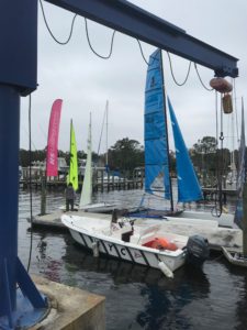 A small white motorboat and colorful sailboats are docked at a marina. Two people are near the boats, and a blue crane structure frames the left side of the image. Trees and more boats are in the background under a cloudy sky.
