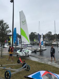People walk near a dock lined with small sailboats on a cloudy day. Flags and sails are visible, along with boat trailers on the grass, and trees and buildings are in the background.