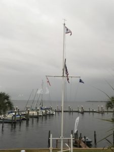A marina on a cloudy day with a tall flagpole displaying multiple flags, boats docked at the piers, and calm water in the background. Palm trees are visible at the edge of the image.