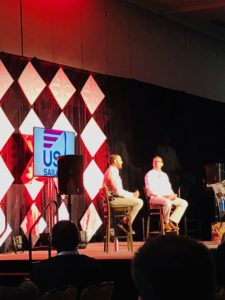 Two men sit on high chairs on a stage, engaging in conversation. Behind them is a patterned backdrop with red and white diamonds and a large screen displaying the "US Sailing" logo. Audience members are visible in the foreground.
