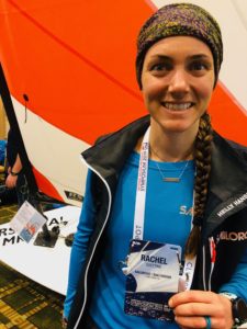 A woman with a braid, wearing a colorful headband and blue shirt, smiles while holding a name badge reading “Rachel Goetting.” She stands in front of a sailboat indoors with an orange and white sail.