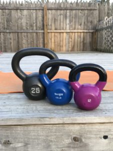 Three kettlebells—black 25 lb, blue 15 lb, and purple 12 lb—are lined up on a wooden deck with an orange exercise mat and a wooden fence in the background.