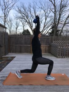A person in athletic wear lunges on an orange mat outdoors, holding a blue kettlebell overhead with both hands. Leafless trees and a wooden fence are visible in the background.