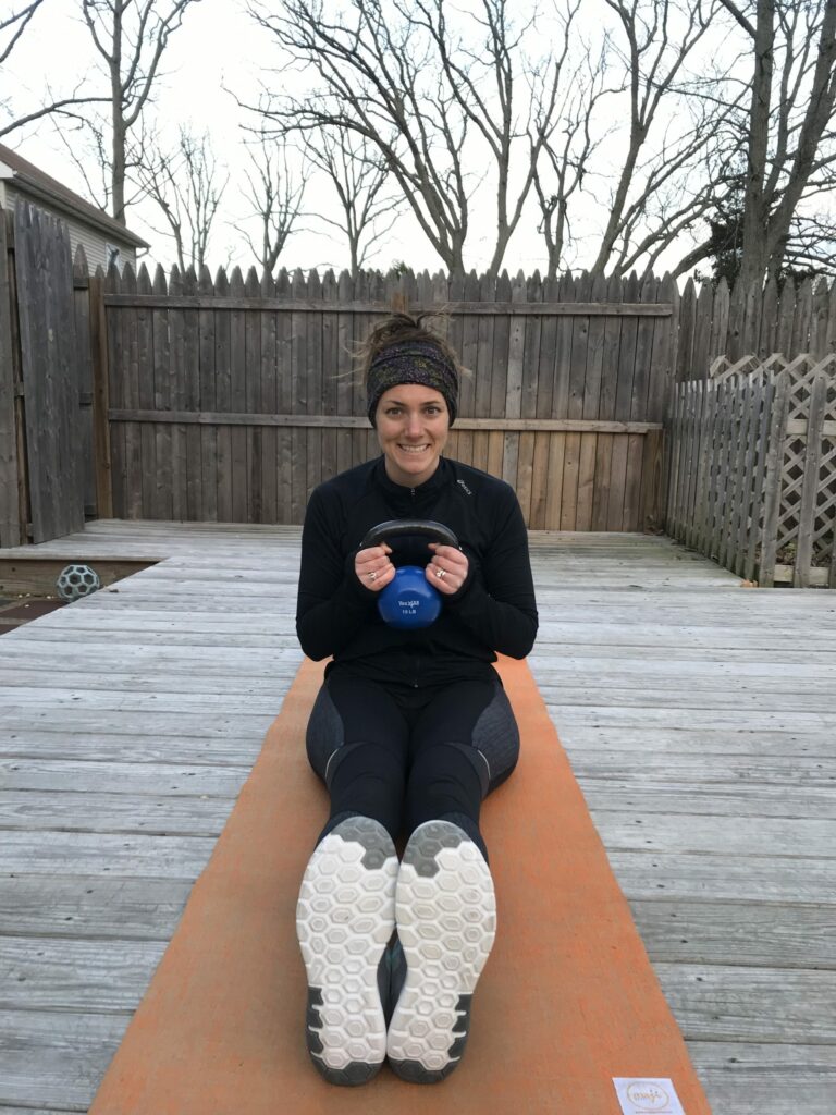 A woman wearing athletic clothes sits on an orange yoga mat outdoors, holding a blue kettlebell, smiling at the camera. Her legs are extended forward and trees without leaves are visible in the background.