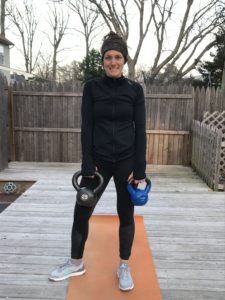 A woman stands on an orange yoga mat outdoors, holding two kettlebells—one black (25 lbs) and one blue. She wears a headband, black jacket, and leggings, and smiles at the camera. Bare trees and a wooden fence are in the background.
