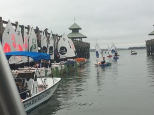 Small sailboats with numbered sails and young sailors gather near a wooden dock, where people are watching. A larger boat labeled "Hilton Head Sailing Center" is in the foreground. The water is calm and overcast.