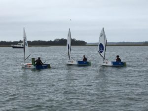 Three small sailboats with single sailors are on the water near the shore, with a larger boat and trees visible in the background under a cloudy sky.