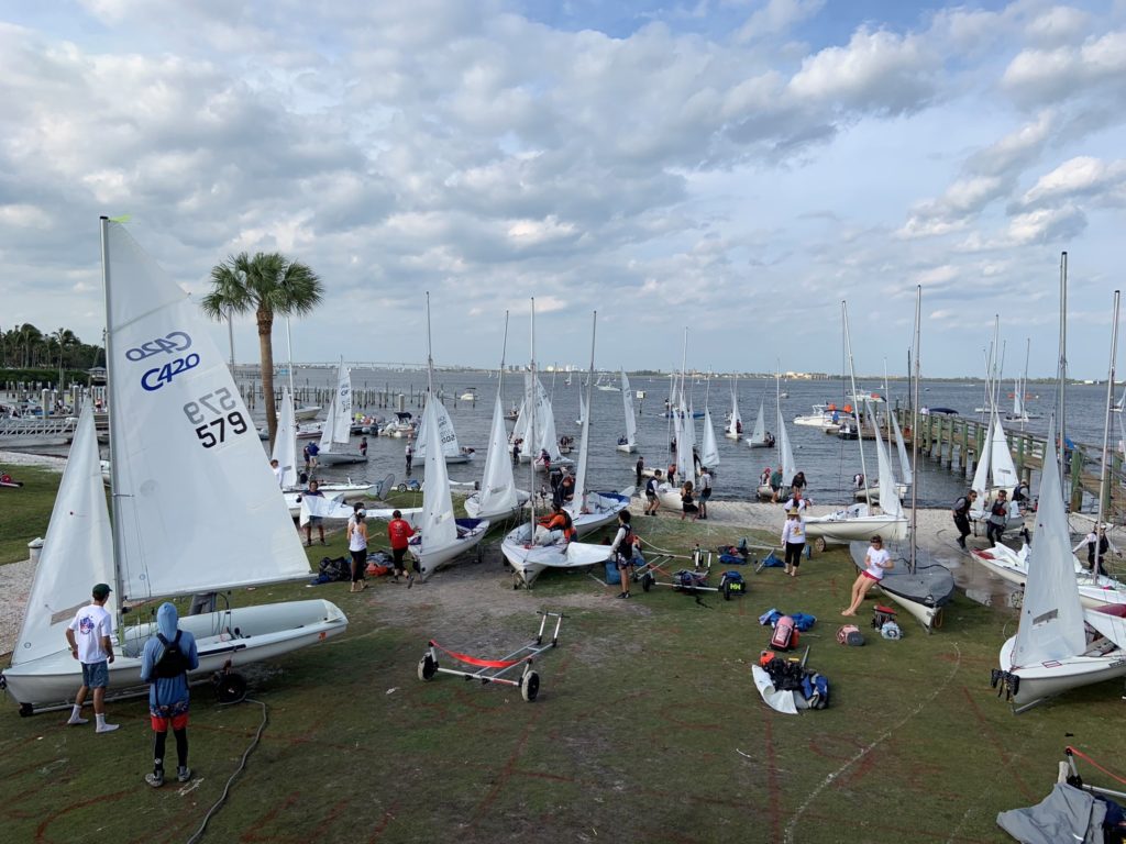 Dozens of people prepare small sailboats on a grassy shoreline next to a lake or bay, with palm trees and docks visible under a partly cloudy sky. Some boats are in the water while others are being worked on.