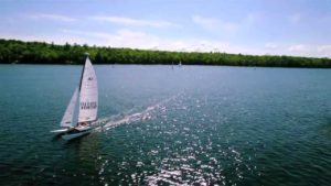 A white sailboat with two people onboard glides across a calm blue lake, leaving a small wake behind. There is a tree-lined shore in the background under a clear, sunny sky.