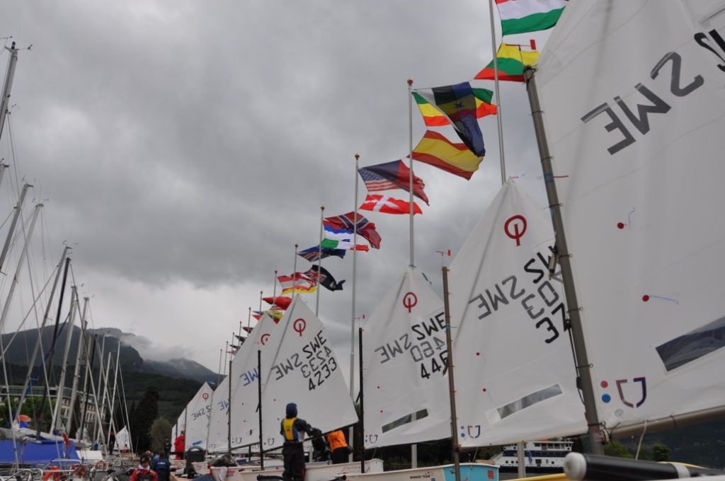A row of sailboats with raised sails and colorful international flags on a cloudy day, docked at a marina with mountains and trees in the background. Several people are standing near the boats.