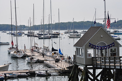 A small pier with sailboats docked, a wooden boathouse with flags, and many sailboats anchored in a calm bay; trees line the distant shore under a hazy sky.